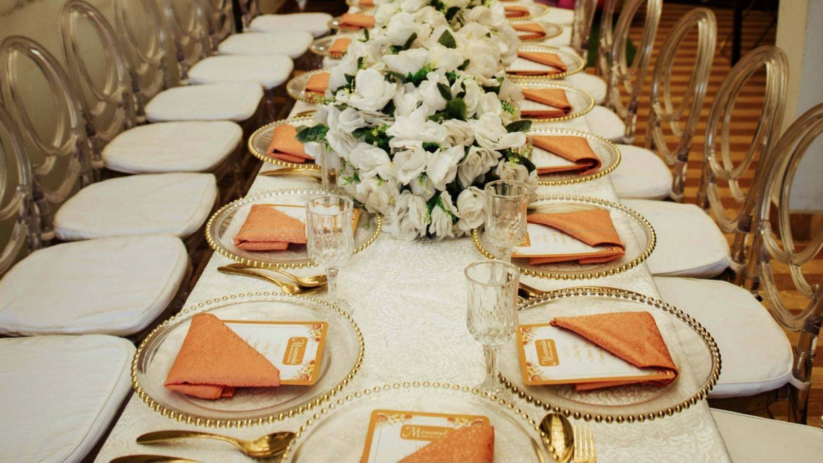 A long celebration table with white roses, gold-rimmed chargers and clear chairs