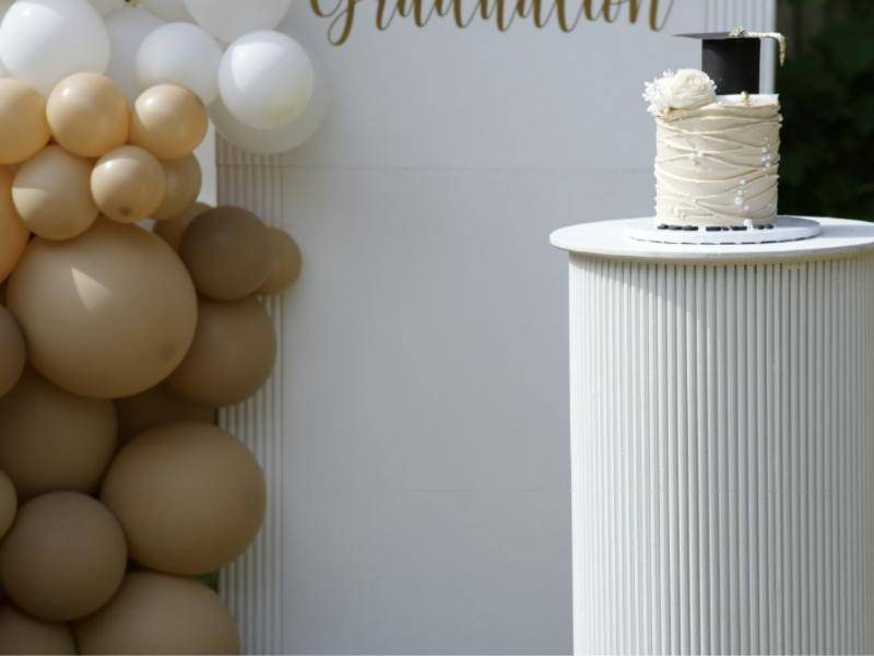 Graduation cake on a white pedestal beside a tan and white balloon arch
